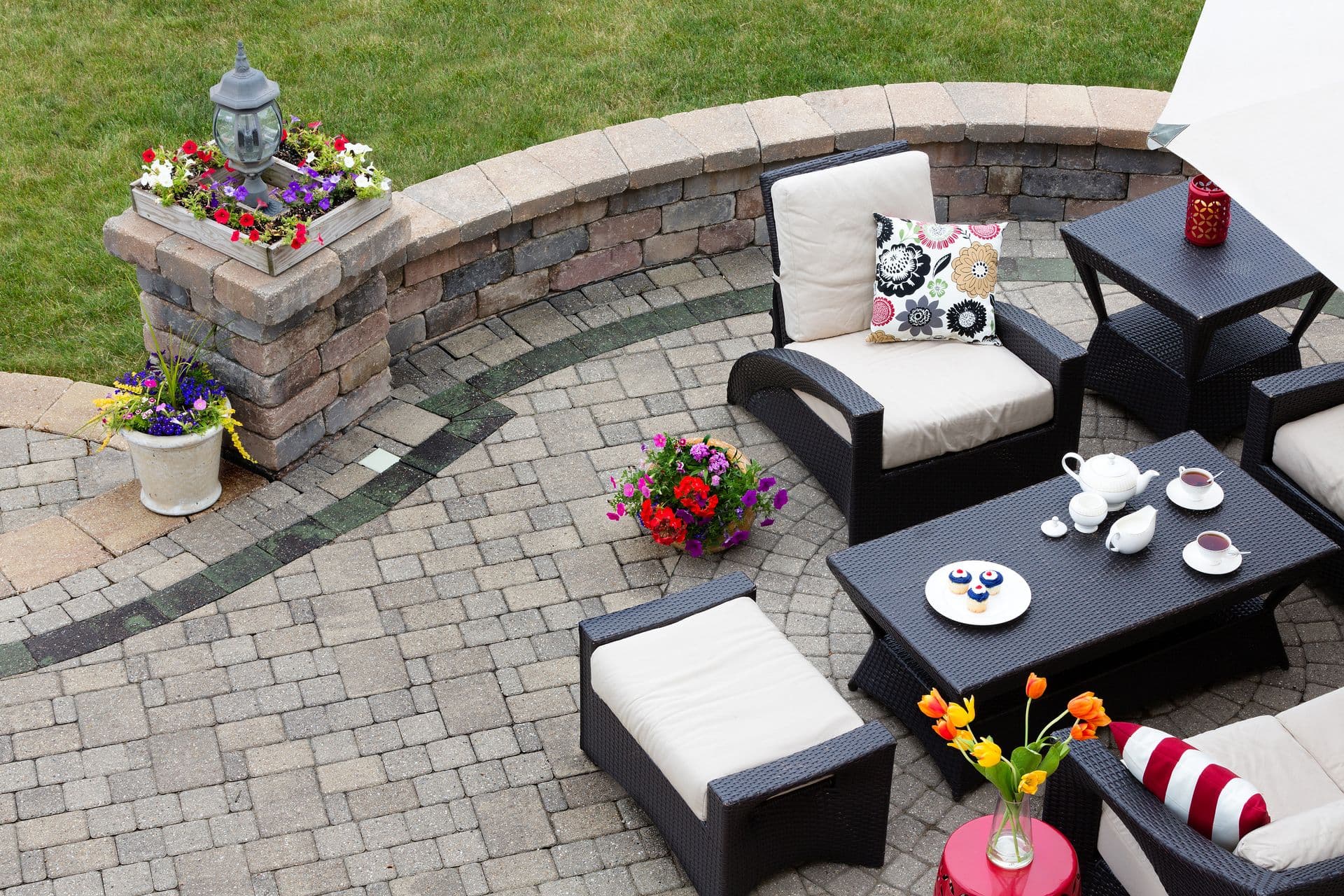 Backyard patio with curved brick seating wall, black wicker conversation set with cream cushions, decorative flowering planters, tea service, and manicured lawn