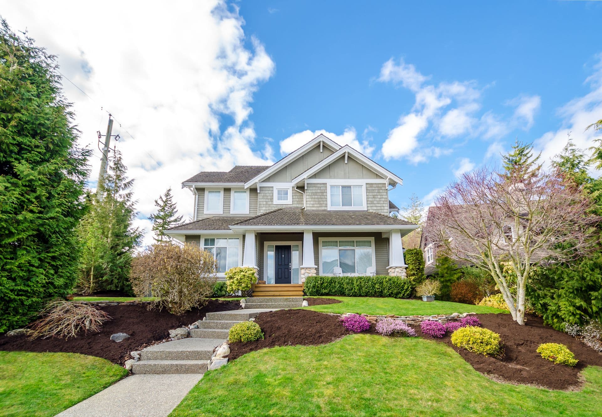 Two-story modern house with white and beige siding, dark roof, and covered front porch, surrounded by manicured lawn, flowering shrubs, and evergreen trees