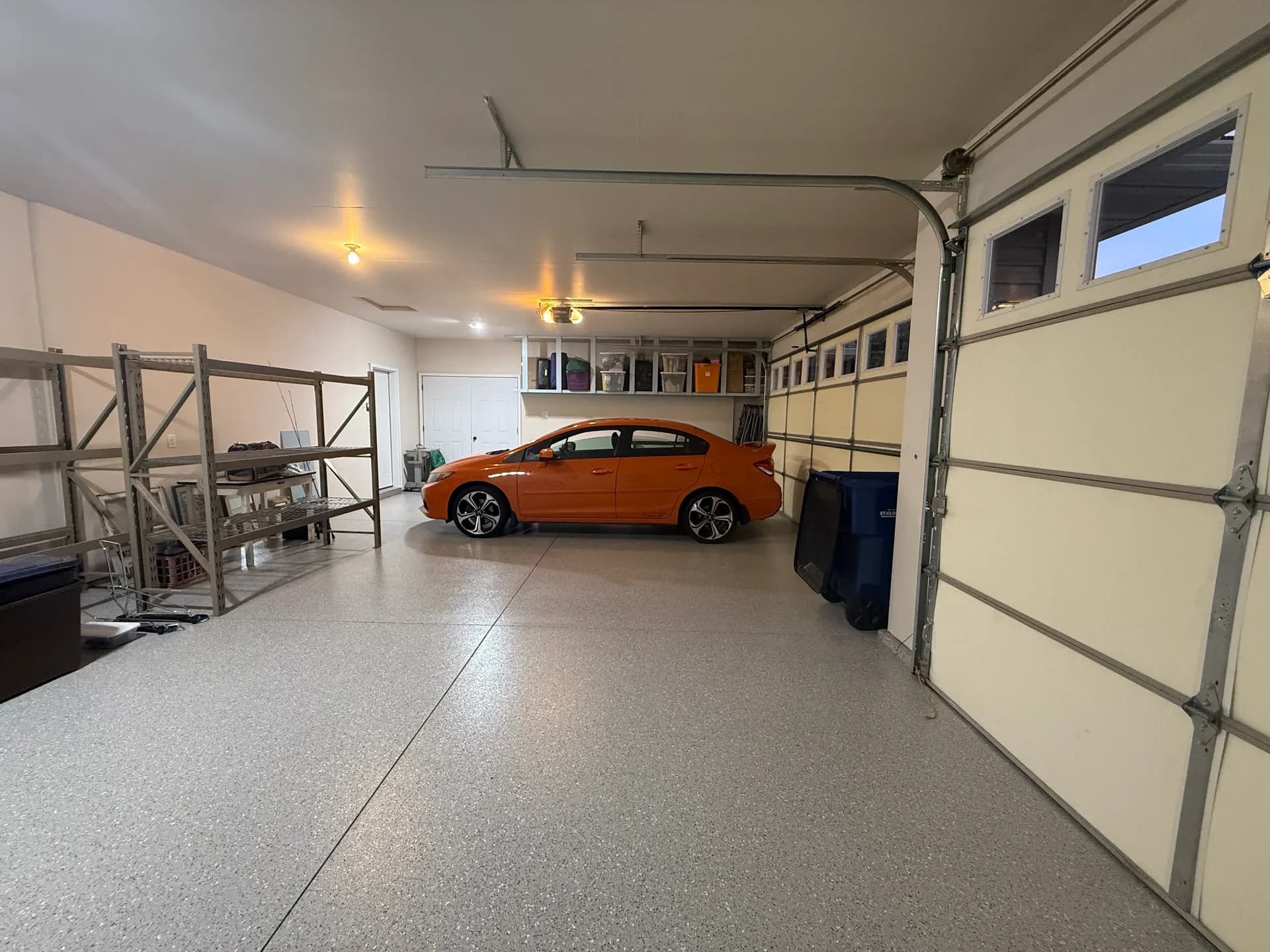 Orange sedan parked in well-organized garage with overhead door open, shelving, and storage racks