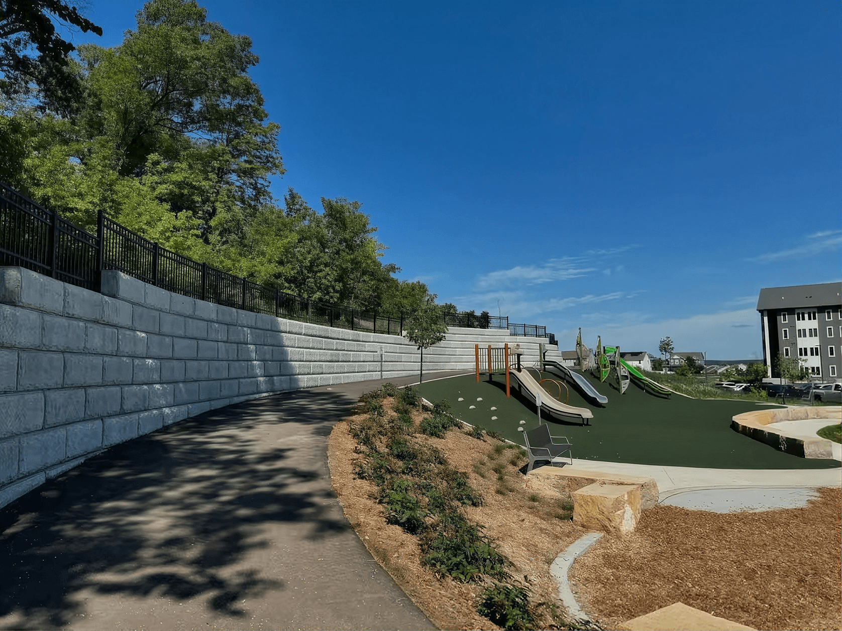 Modern playground with green artificial turf, slides, climbing equipment, and sandbox bordered by concrete retaining wall and black railing on sunny day