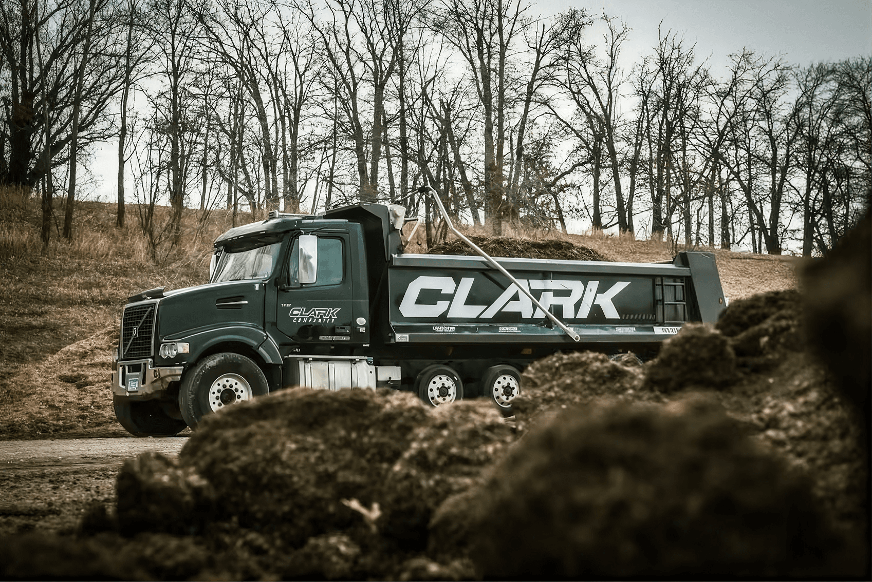 Clark dump truck loaded with mulch at a construction site surrounded by bare trees and soil piles