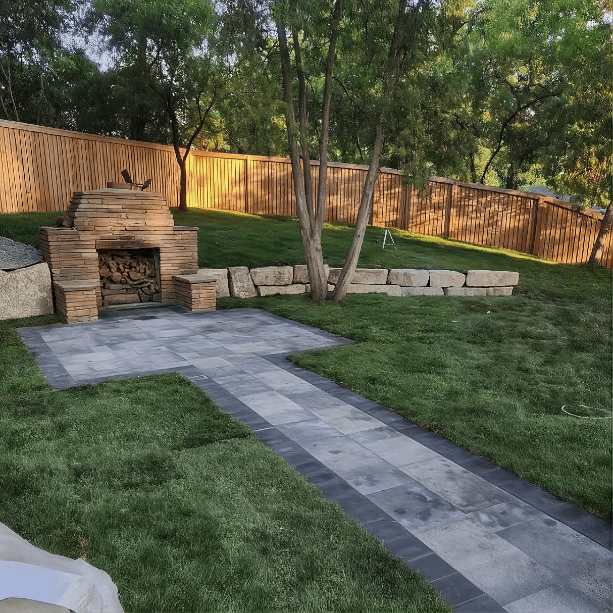Backyard patio area with stone fireplace, paver pathways, green lawn, wooden deck seating, mature trees, and metal fence with warm lighting at dusk