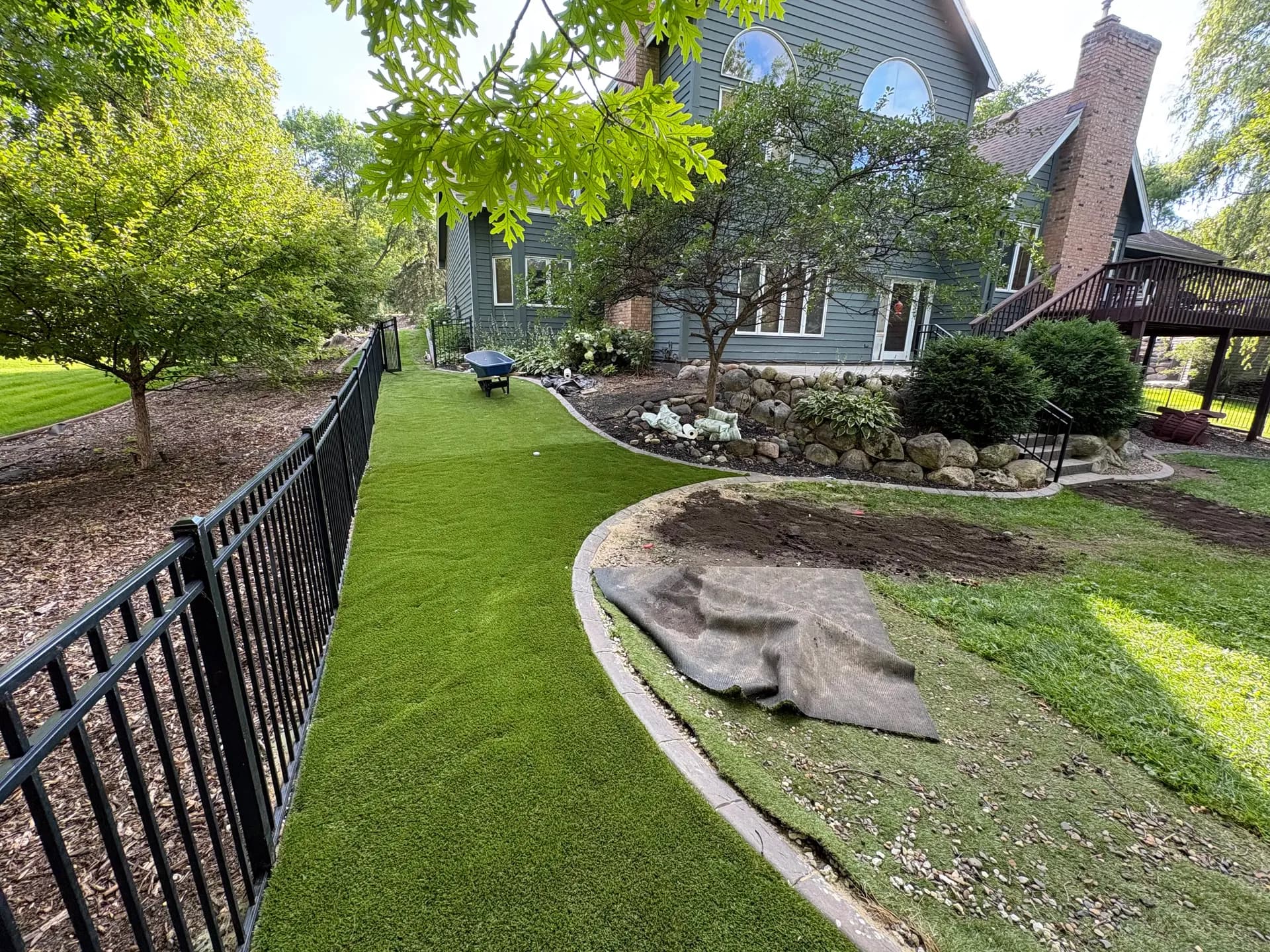 Residential backyard with artificial turf, stone landscaping features, black metal fencing, brick house with deck, and mature shade trees surrounding the property